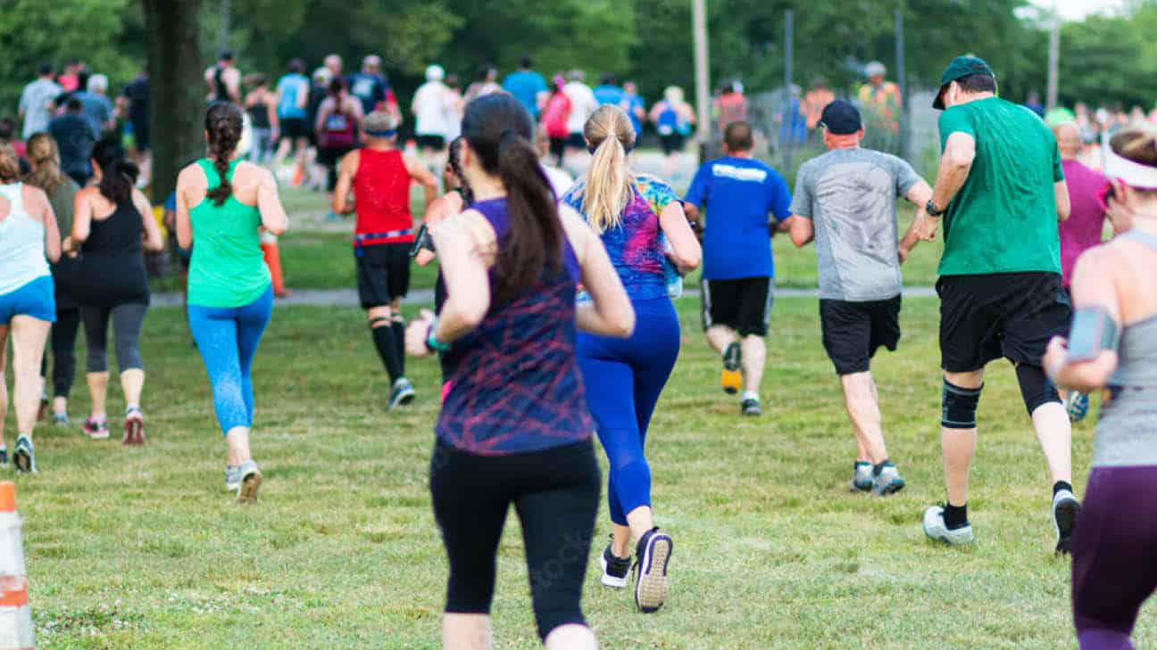 Group of people running outdoors in a 5K race, wearing athletic clothes in various colors, on a grassy course surrounded by trees.